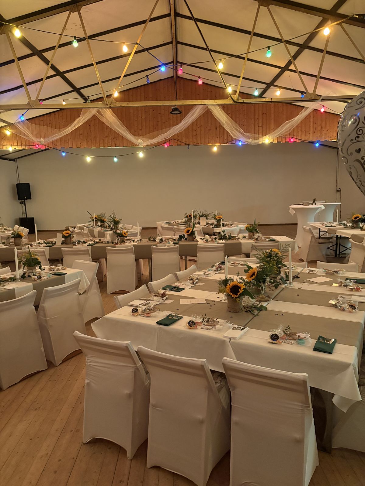 Festlich eingedeckter Saal mit weißen Stuhlhussen, Sonnenblumen-Deko und Lichterketten